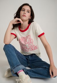 T-shirt crème avec col et manches rouges, arborant le texte "Welcome to the Upside Down" en rose, associé à un jean bleu et des baskets blanches.