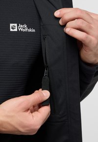 Giacca nera con tasca con cerniera. Il materiale presenta un aspetto texturizzato con una finitura liscia. Logo di Jack Wolfskin visibile.