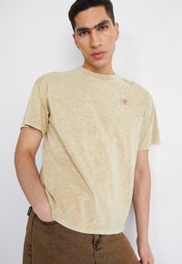 Camiseta de algodón beige claro con un acabado texturizado de lavado mineral. Presenta un pequeño parche con logo en el pecho y mangas cortas.