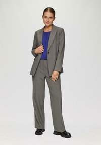 Grijze op maat gemaakte suit met een enkele knoop, gecombineerd met een donkerblauwe blouse, met rechte broekspijpen en glanzende zwarte schoenen.