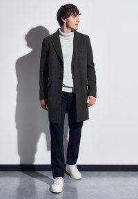 Tmavě šedý vlněný kabát s přiléhavým střihem, jednoduchým zapínáním, klopami a dvěma postranními kapsami, nošený přes světle šedý rolák.