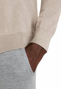 Maglione beige a maglia con polsini a coste, abbinato a pantaloni grigi chiari con texture e una tasca. Tessuto morbido e dettagli di design puliti.