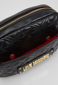 Svart kviltad konstläderväska med guldfärgad hårdvara, med en röd insida och en utsida med guld "LOVE MOSCHINO" logoplåt. Dragkedjestängning.