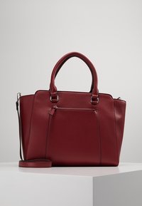 Czerwona skórzana torebka z dwoma uchwytami, odpinanym paskiem na ramię, przednią kieszenią na suwak oraz wyraźnie zarysowanym, prostokątnym kształtem. Gładka faktura i minimalna liczba szwów.