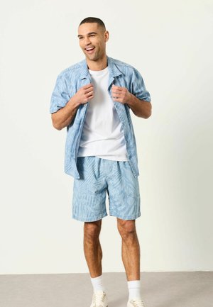 Mand smiler og justerer kortærmet blå mønstret skjorte over en hvid T-shirt, iført matchende shorts, hvide sokker og sneakers.