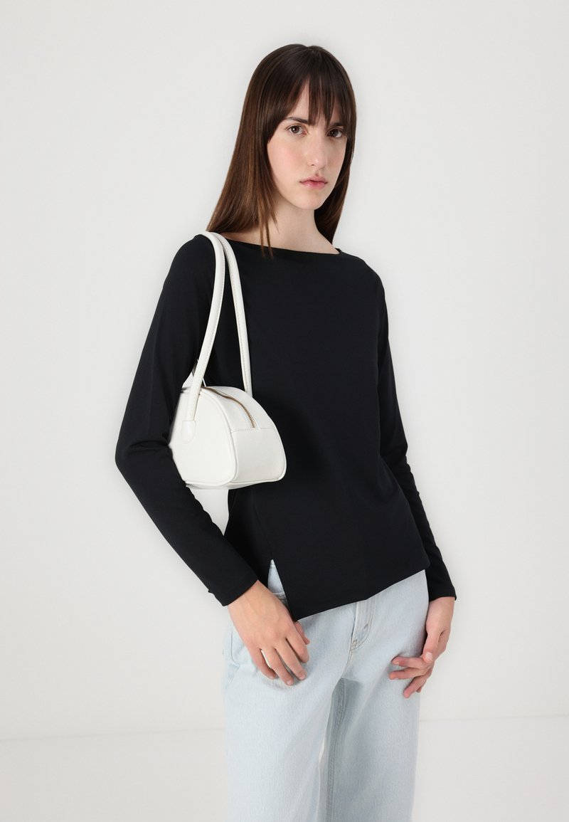 Schwarzes Langarmoberteil mit einem Bootsausschnitt, kombiniert mit hellblauen weiten Jeans. Hält eine kleine weiße Handtasche in runder Form mit Reißverschluss.