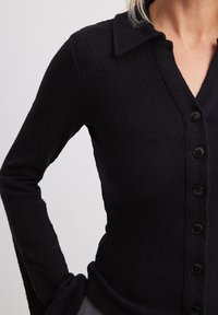 Zwarte cardigan van gestructureerde stof met een kraaghalslijn en een knoopsluiting, lange mouwen en een getailleerd silhouet.