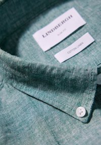 Col de chemise en lin coton vert avec boutons blancs et étiquettes en tissu blanc à l'intérieur indiquant la marque et le matériau.