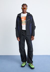 Black and navy track jacket with hood, paired with matching pants. White T-shirt with orange Nike logo. Neon green shoes with black accents.