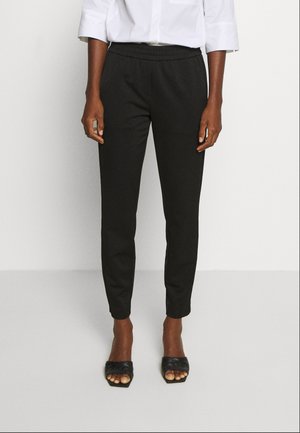 Person iført sorte slim-fit bukser med elastisk talje, hvid langærmet skjorte og sorte åbne hælede sandaler.
