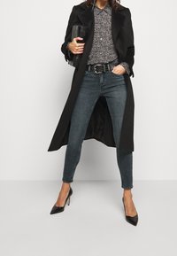 Abrigo largo negro sobre una camisa gris con estampado, con jeans skinny azul oscuro y tacones negros de punta. Clutch negro con un acabado texturizado.