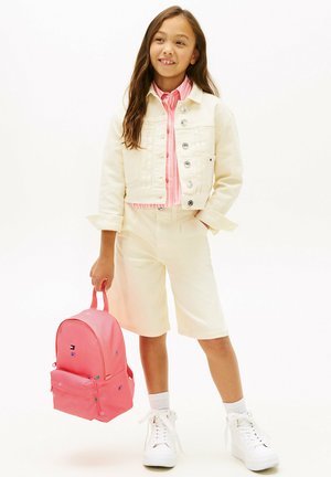 Niña con chaqueta crema y pantalones cortos, con camiseta de rayas rosas, sosteniendo una mochila rosa, de pie sobre un fondo blanco.