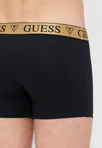 Melni boksa šortai ar zelta elastīgu jostu, uz kuras ir melnais "GUESS" logo un emblēma. Izgatavoti no gludas, elastīgas auduma.
