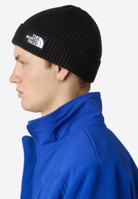 Căciulă din tricot negru cu carouri, dotată cu o etichetă albă, purtată cu o geacă de fleece albastru strălucitor, având un guler înalt și texturi contrastante.