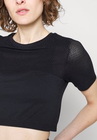 Top cropped preto com mangas curtas, apresentando um painel em rede nos ombros e um corpo de tecido liso e elástico.