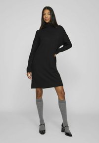 Black knit dress with a high neckline and long sleeves. Paired with grey knee-high socks and pointed black heels. Soft texture and relaxed fit.