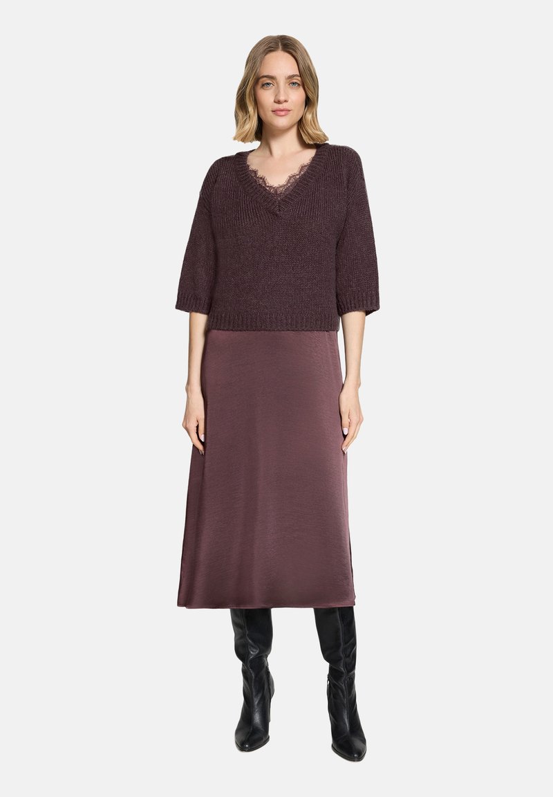 Femme portant un pull en maille violet foncé avec bordure en dentelle, une jupe midi assortie et des bottes noires montantes, debout devant un fond blanc.