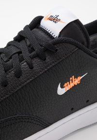 Svart lädersneaker med texturerad yta, kontrasterande vit och orange Nike-logotyp, samt svarta snören. Vit gummisula med synliga sömmar.