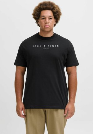 Muškarac s kovrčavom kosom i pjegama nosi crnu majicu "Jack & Jones Premium" i bež hlače, stoji naslonjen na jednostavnu bijelu pozadinu.