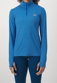 Žena na sobě má modré sportovní tričko s dlouhým rukávem a předním zipem, doplněné o odpovídající modré legíny.