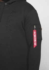 Crna dukserica s reljefnim logotipom "ALPHA INDUSTRIES", džepom s patent zatvaračem na rukavu i crvenom oznakom "REMOVE BEFORE FLIGHT".