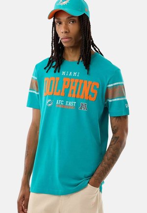 Uomo con i capelli lunghi che indossa un cappellino teal dei Miami Dolphins e una maglietta coordinata con scritta arancione e pantaloni beige, in piedi con una mano in tasca.