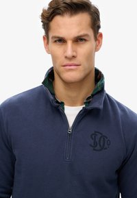 Pulóver azul marino con cuello de media cremallera, que presenta una camiseta de cuadros verde debajo. Logotipo negro en el pecho, fabricado en una mezcla de algodón.