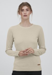 Beige neulepaita, jossa on teksturoitu hunajakennokuvio, klassinen pyöreä pääntie, resorit hihoissa ja helmaresorissa sekä pieni brändilappu alareunassa.