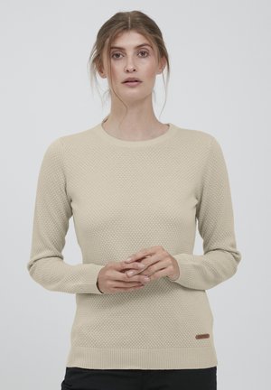 Beige strikkegenser med teksturert bikake-mønster, klassisk rund hals, ribbestrikkede mansjetter og kant, med en liten merkelapp nederst.