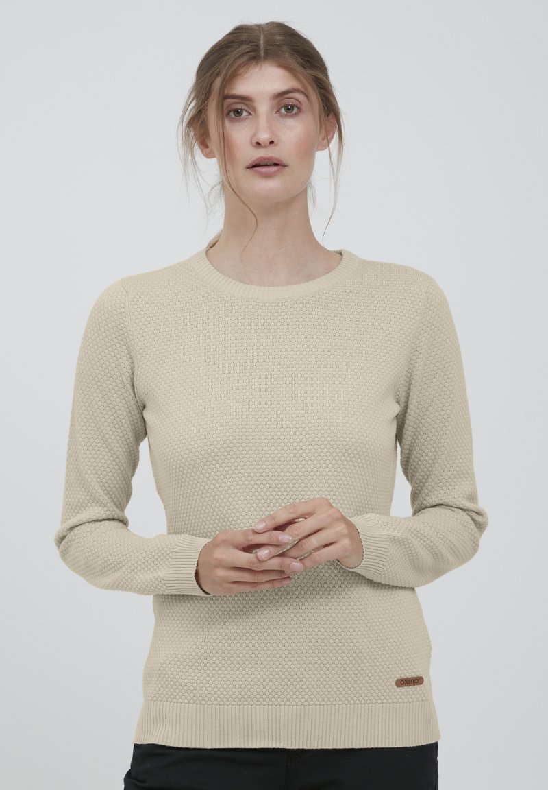 Beige neulepaita, jossa on teksturoitu hunajakennokuvio, klassinen pyöreä pääntie, resorit hihoissa ja helmaresorissa sekä pieni brändilappu alareunassa.
