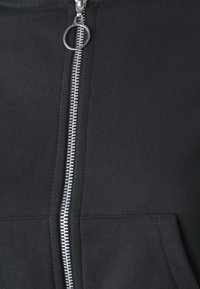 Veste à capuche noire zippée avec une fermeture éclair en métal argenté munie d'une tirette circulaire, tissu texturé et détail d'une poche avant.
