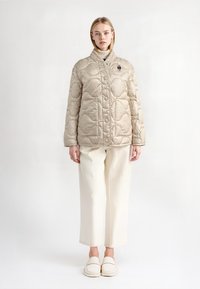 Beige gewatteerde pufferjack met een hoge kraag, voorzien van een drukknoopsluiting en een textuurpatroon, gedragen met witte wijde broekspijpen en instappers.