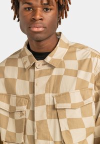 Beige ruudullinen paita, jossa on kaksi etutaskua, napitus ja teksturoitu kangas. Vasemmassa taskussa on logo.