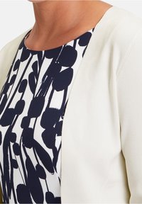 Vit kofta med rund halsringning över en marinblå och vit blommönstrad topp. Koftan har en slät textur och en minimalistisk design.