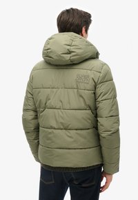 Olivengrøn pufferjakke med hætte, quiltet design og broderet logo på ryggen. Tekstureret bundkant, kombineret med mørke jeans.