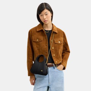 Žena má na sebe hnedú semišovú bundu cez čierne tričko a svetlomodré rifle, nesie malú čiernu kabelku so zlatým reťazovým remienkom.