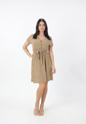 Vestido beige de manga corta y escote en V con botones y cintura ajustada, con un corte holgado y textura suave, combinado con tacones transparentes.
