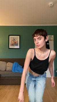 Svart crop top med tynne stropper, paret med lyseblå høyt livet jeans. Naturlig tregulv, grønn vegg og rammet blomsterkunstverk i bakgrunnen.