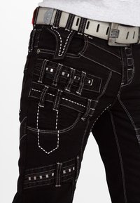 Pantaloni in denim nero con molteplici tasche utilitarie e dettagli in cucitura bianca. Include una cintura grigia e passanti per cintura a motivo, enfatizzando la funzionalità.