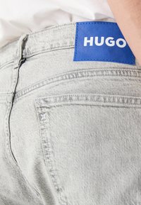 Šviesiai pilki džinsai su mėlynu odiniu ženkliuku, pažymėtu "HUGO", ant juosmens. Džinsai turi matomus siūlus ir minimalistinio dizaino detales.