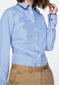 Camisa de cuadros vichy azul y blanca con cuello, cierre de botones en la parte delantera y ribete negro en los puños y el cuello. Combinada con pantalones color caqui.