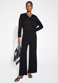 Jumpsuit nero con scollo a V, maniche lunghe e taglio a gamba larga. Realizzato in tessuto testurizzato con una leggera lucentezza. Accessorio: borsa metallica argento.