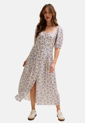 Frau trägt ein weißes Midikleid mit blauem Blumenmuster, kurzen Puffärmeln, einem vorderen Schlitz und weißen Turnschuhen vor einem einfarbigen Hintergrund.