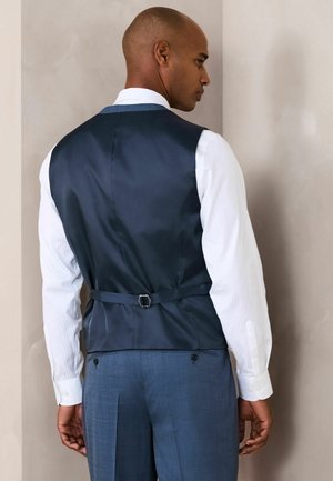 Mann trägt eine marineblaue Weste mit verstellbarem Schnallenverschluss über einem weißen Hemd und blauen Hosen, steht vor einer schlichten Wand und blickt weg.