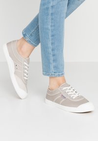 Beige canvas sneakers with white rubber soles, featuring lace-up design, side eyelet details, and a label on the side.