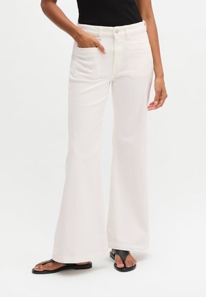 Osoba w białych szerokich spodniach z wysokim stanem, czarnym bezrękawniku i czarnych sandałach z odkrytymi palcami, ręka częściowo w kieszeni.