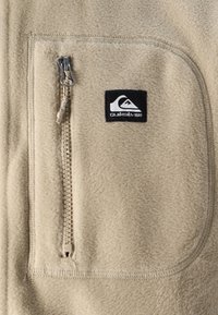 Beige fleece jacket with zippered chest pocket, textured fabric, and Quiksilver logo patch. Visible stitching and smooth zipper detail.
