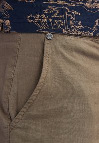 Pantaloni in lino color sabbia con tasca frontale, dotati di chiusura a bottone metallico e cuciture a vista. Sullo sfondo si intravede una parte superiore con un motivo scuro.
