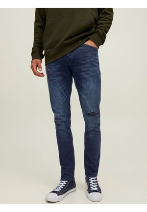Jeans de mezclilla azul oscuro con acentos desgastados, de corte ajustado, con dos bolsillos frontales, combinados con zapatillas blancas y una sudadera oliva.