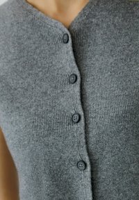 Grauer Strickcardigan mit V-Ausschnitt, der fünf schwarze Knöpfe auf der Vorderseite und eine gerippte Textur aufweist. Das Material scheint weich und gemütlich zu sein.